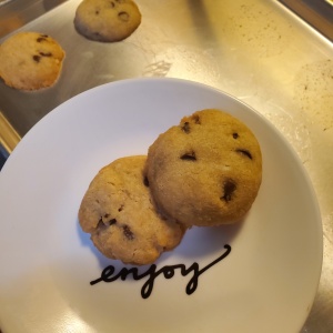 Two chocolate chip cookies arranged on a dessert plate emblazoned with the word ENJOY. In the background, two additional cookies remain on a baking sheet
